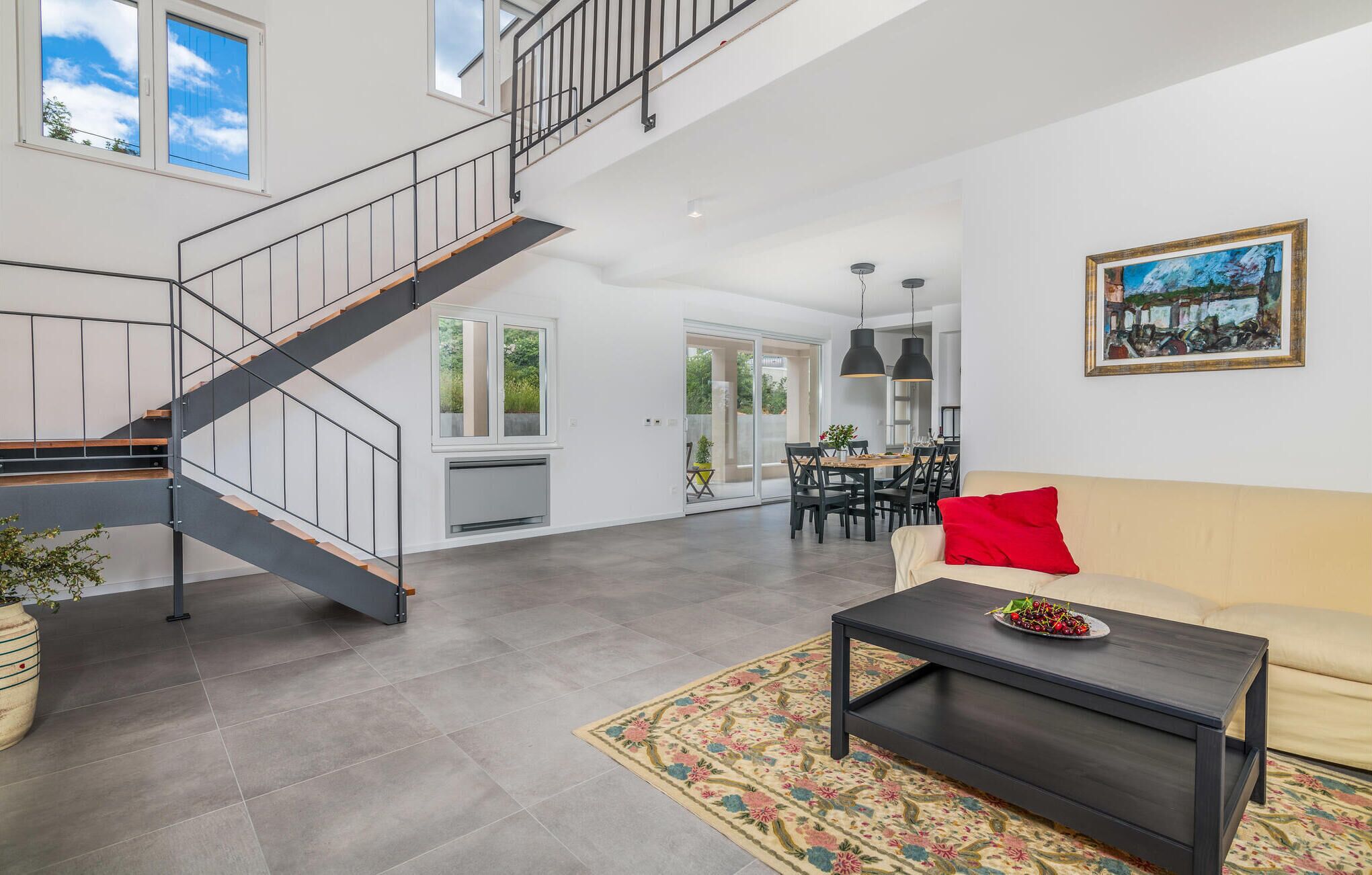 A spacious living area with a staircase, a dining area, and a sofa with a red pillow.