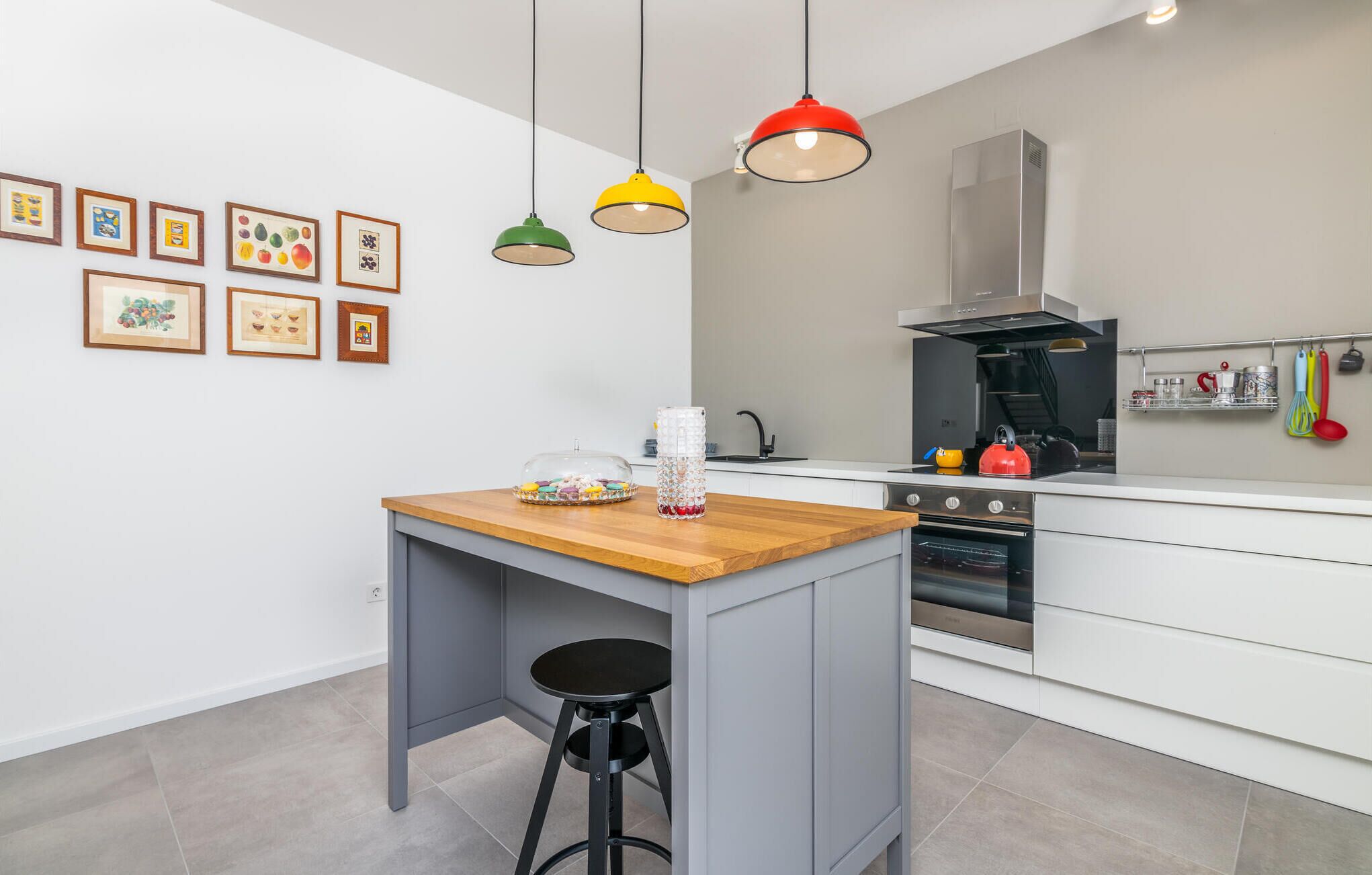 A modern kitchen with white cabinets, a wooden island, and a black stool. There are colorful pendant lights, a stainless steel range hood, and a microwave.