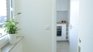 A narrow hallway with a potted plant, a window, and a view into a kitchen area with cabinets and appliances. - Fresh New EU COMMISSION area apartment  (Bruxelles)