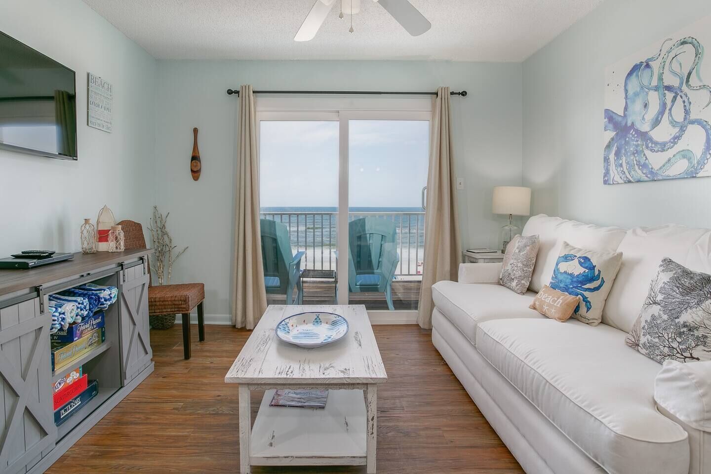 A living room with a white sofa, a coffee table, a TV, and a balcony with beach chairs.