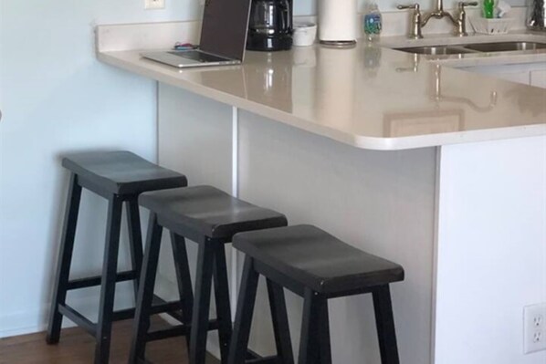 A kitchen with a white countertop, a coffee maker, and three black stools. - Fall Sale! Aamazing View! 2BR/1B Gulf Front! (Gulf Shores)