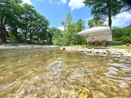 A clear river with rocks, a tent with chairs, and trees in the background.