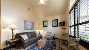 A living room with a brown leather sofa, a coffee table, a dining table with chairs, a TV mounted on the wall, and large windows with blinds.