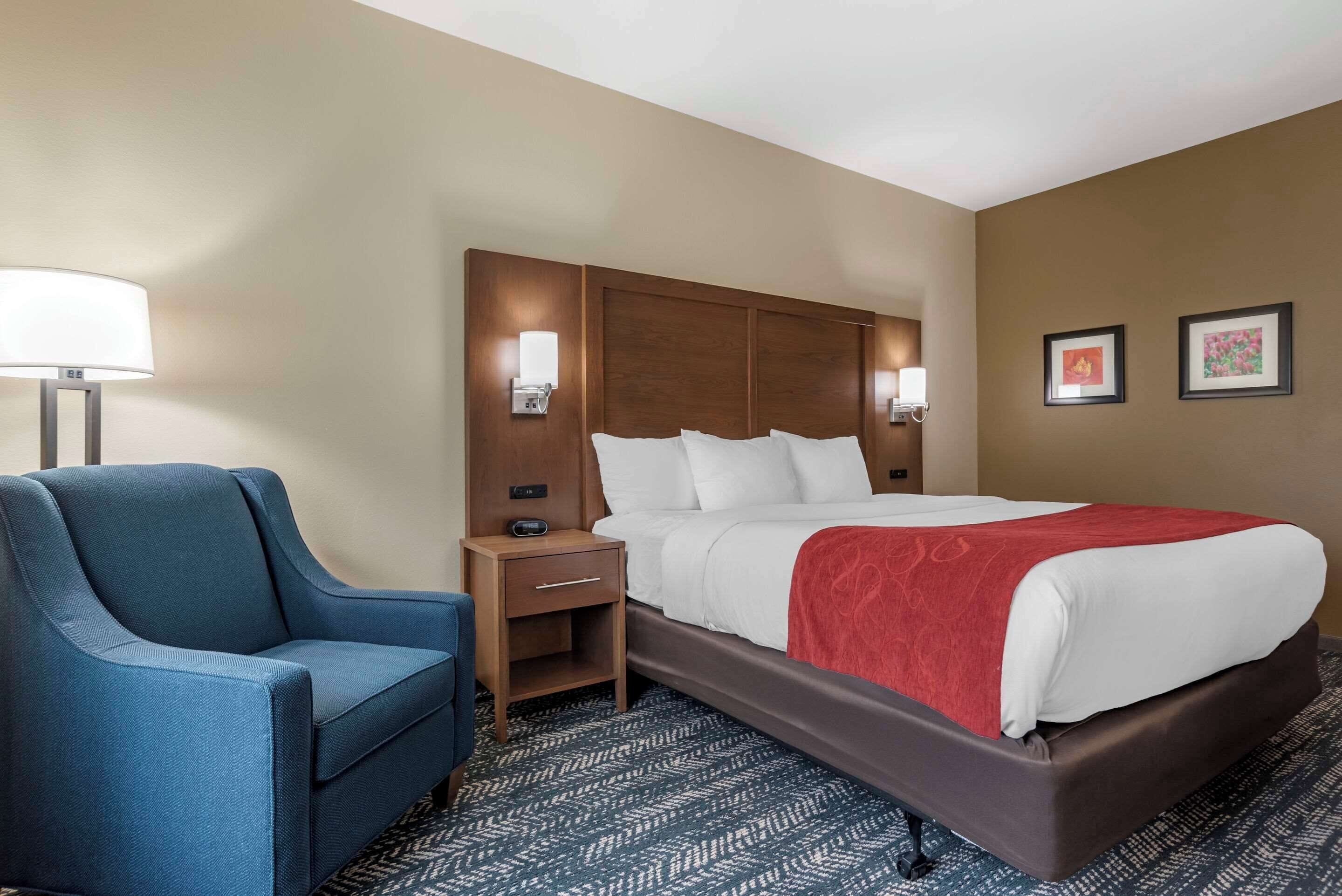 Habitación de hotel con una cama, un sillón azul, una mesita de noche, una lámpara y dos cuadros en la pared.