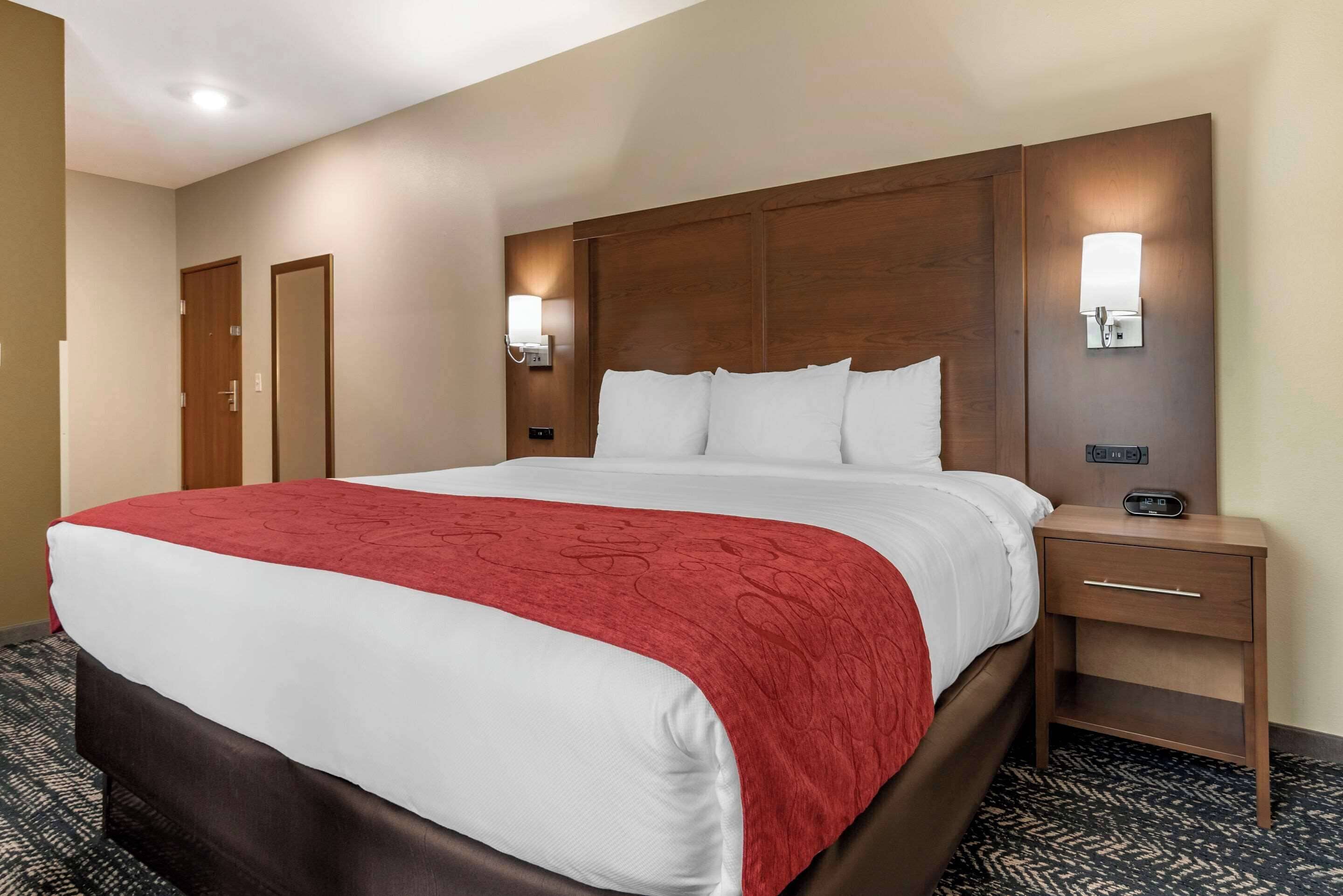 Habitación de hotel con una cama grande, cabecera de madera, dos mesitas de noche con lámparas y suelo alfombrado.