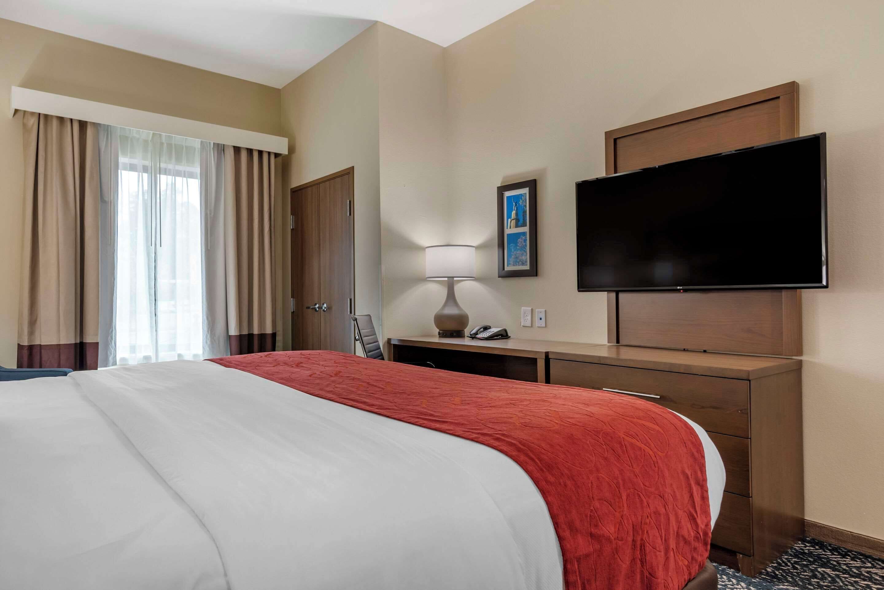 A hotel room with a large bed, a desk with a lamp, a flat-screen TV, and a door.