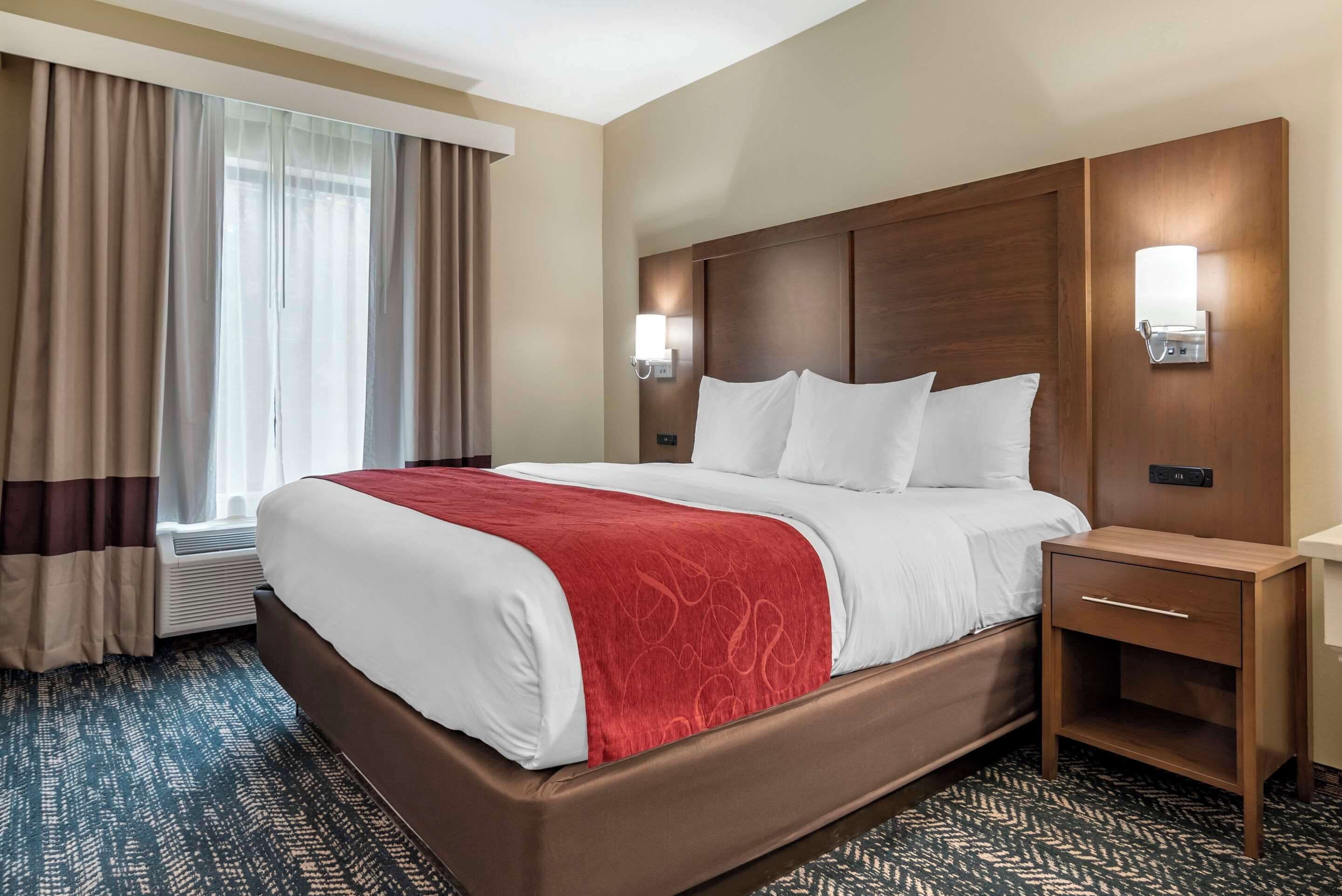 A hotel room with a large bed, a wooden headboard, two bedside lamps, a nightstand, and a window with curtains.