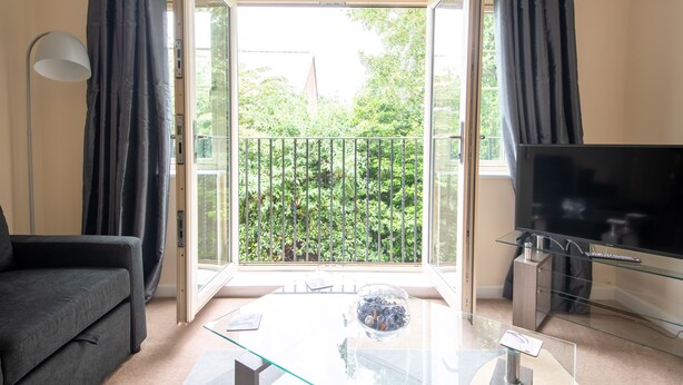 A living room with a sofa, a glass coffee table, a TV on a glass stand, and a balcony with a view of greenery. - No18 (Mansfield)