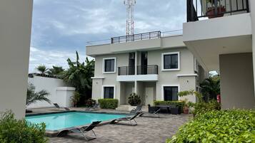 A modern building with a swimming pool, lounge chairs, and potted plants.