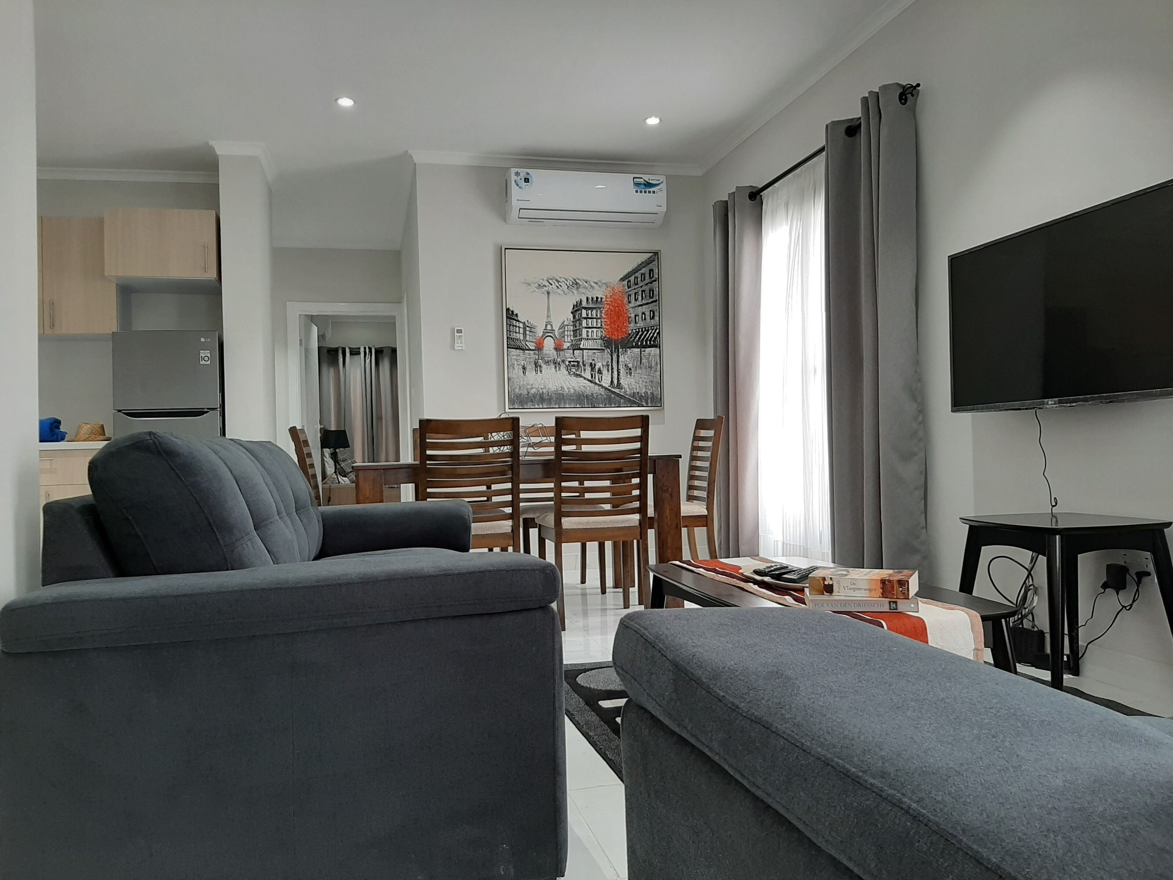 Een moderne woonkamer met een grijze bank, een eethoek met houten stoelen, een flatscreen televisie en een airconditioner aan de muur.