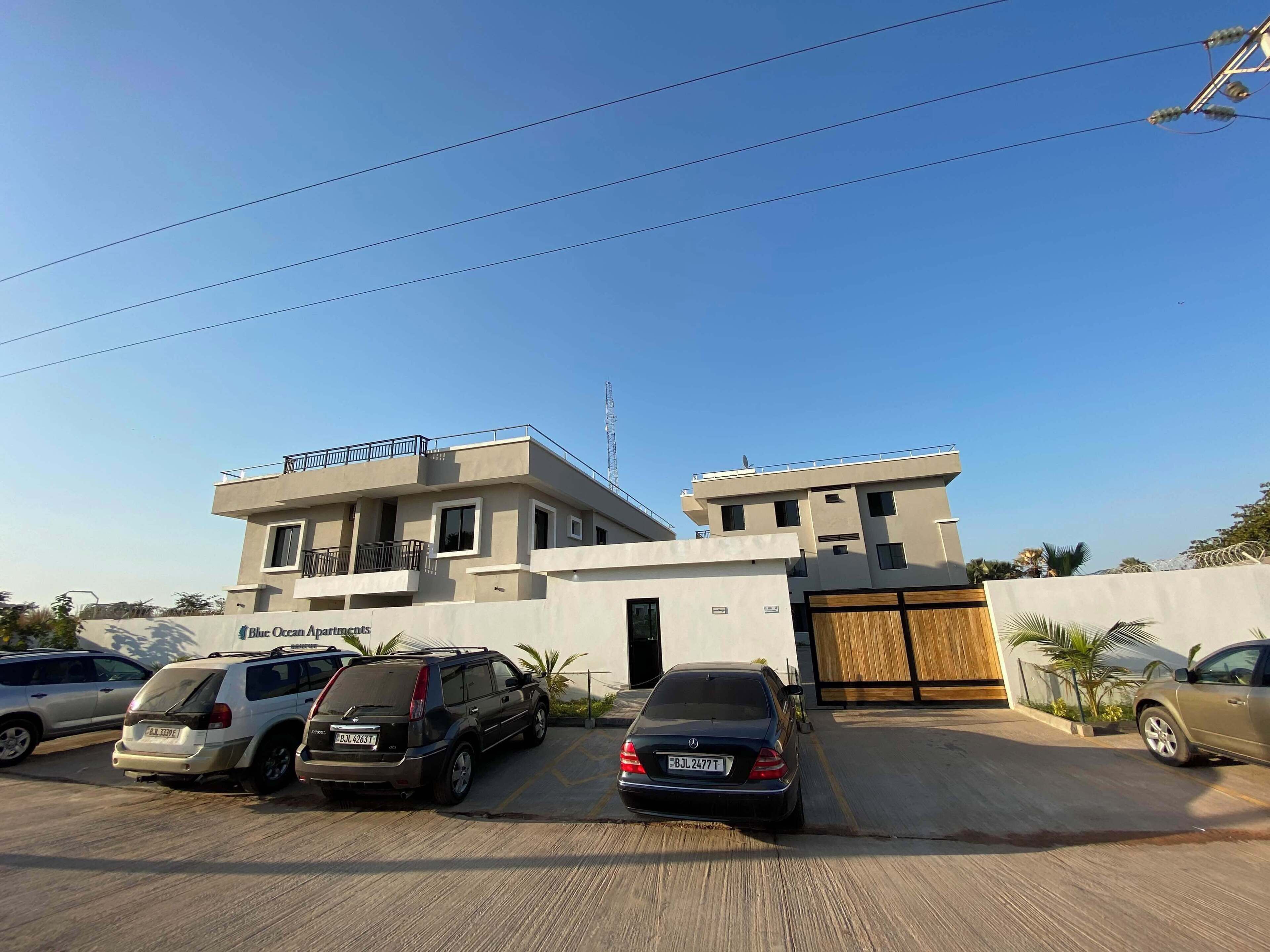 A modern building with a flat roof and a gated entrance. There are several parked cars in front.