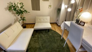 Ett litet, ombonat rum med en vit soffa, ett litet bord och en grön matta. På bordet står en växt, en inramad tavla och en lampa.