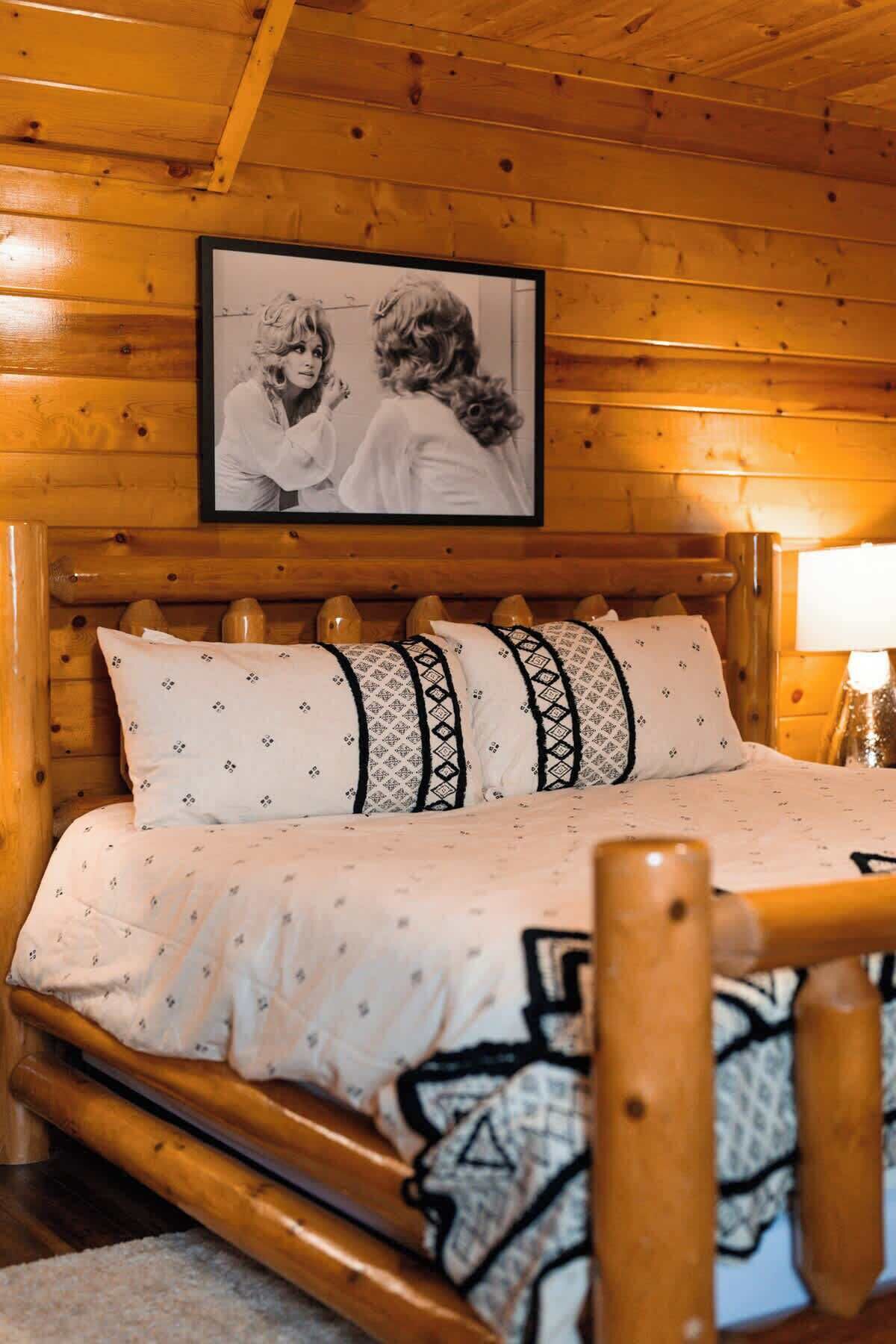 A wooden cabin bedroom with a bed, pillows, a framed picture on the wall, and a lamp.