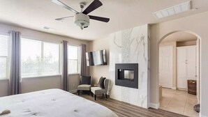 A bedroom with a large bed, a ceiling fan, a flat-screen TV mounted on the wall, and a view of the hallway.