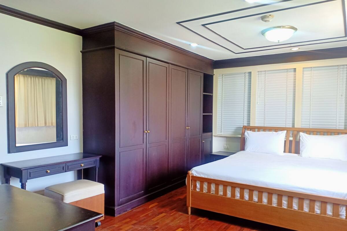 A bedroom with a large wardrobe, a desk, a bed, and a window with blinds.