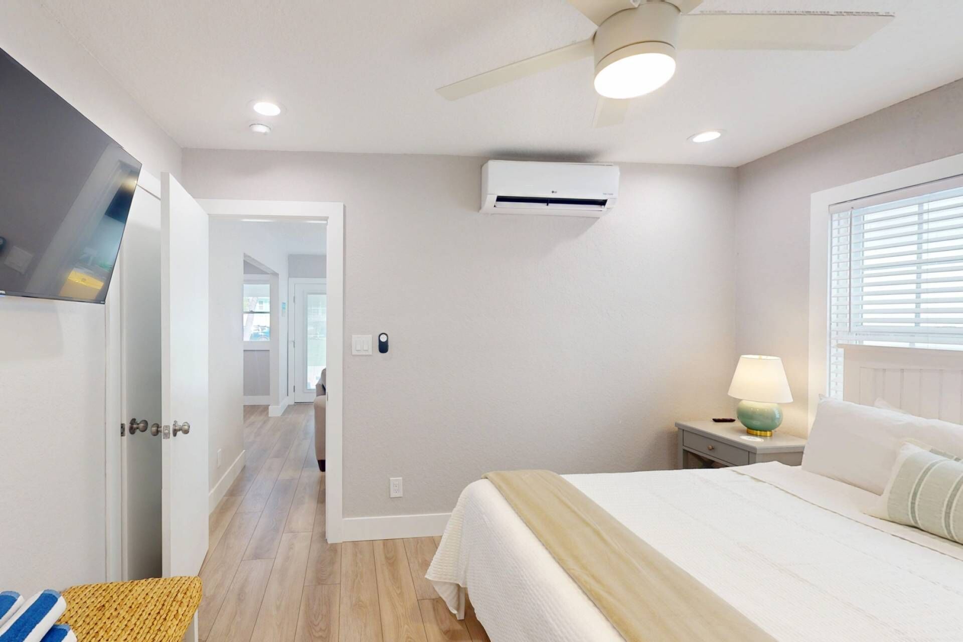 A bedroom with a bed, a nightstand, a lamp, an air conditioning unit, a ceiling fan, and a TV mounted on the wall.