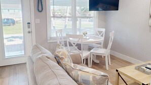 A living room with a white sofa, a round dining table with white chairs, a TV mounted on the wall, and a door with a window.