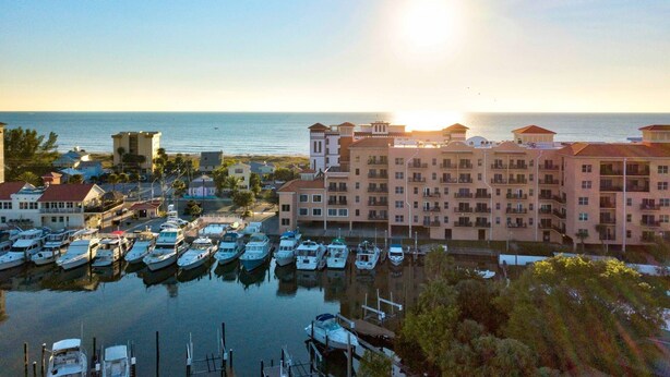 A marina with multiple boats docked, surrounded by multi-story buildings with balconies, and a clear view of the ocean.