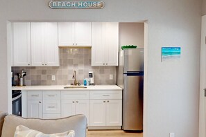A compact kitchen with white cabinets, a stainless steel refrigerator, and a sink. A sign above the cabinets reads "BEACH HOUSE."