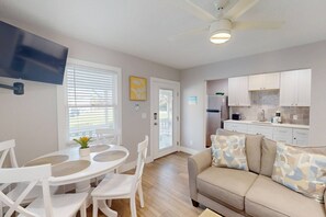 A modern kitchen with white cabinets, a built-in oven, and a refrigerator. There is a dining area with a white table and chairs, and a beige sofa with patterned cushions.