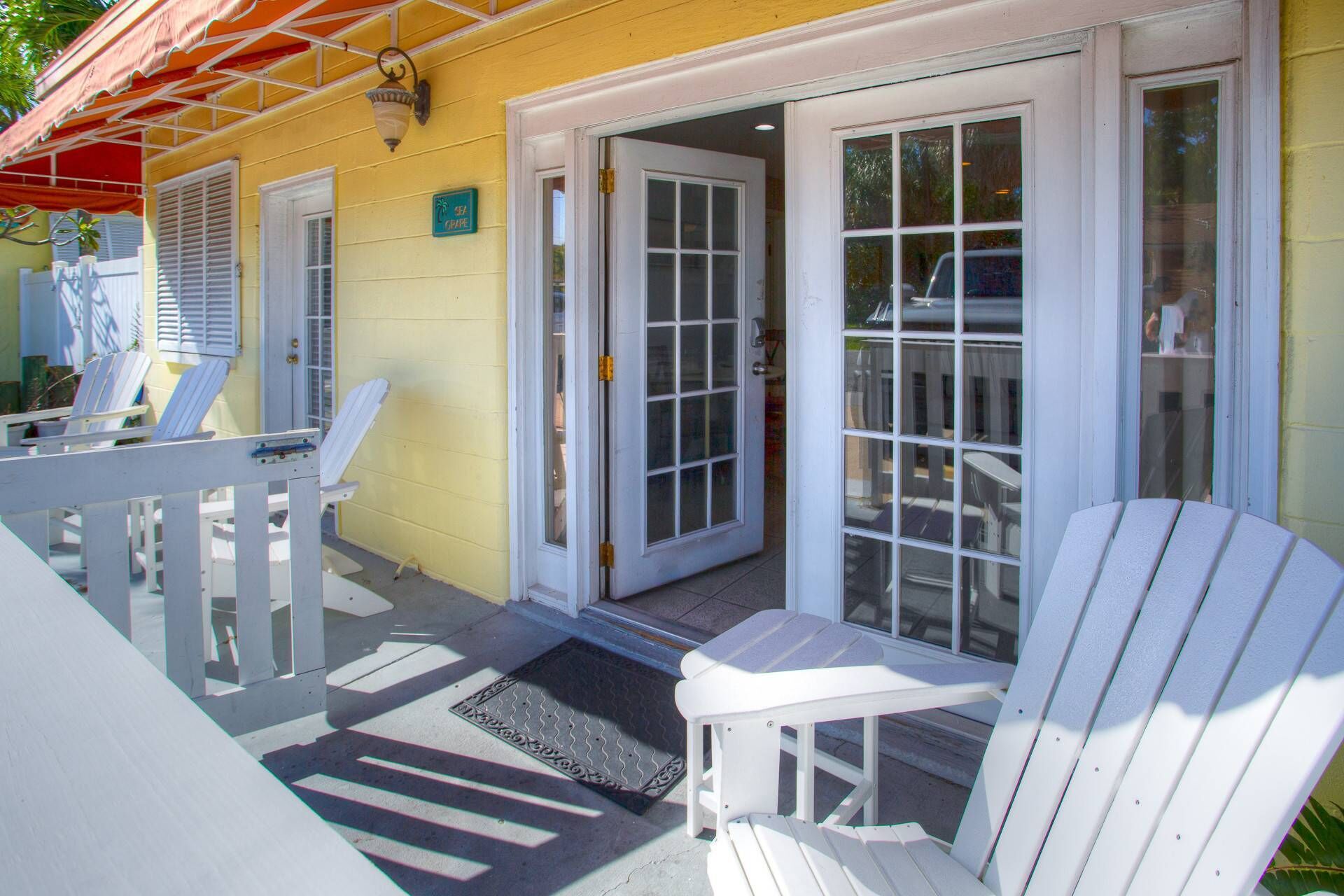 A yellow building with white doors and windows, outdoor seating with white chairs, and a small table.