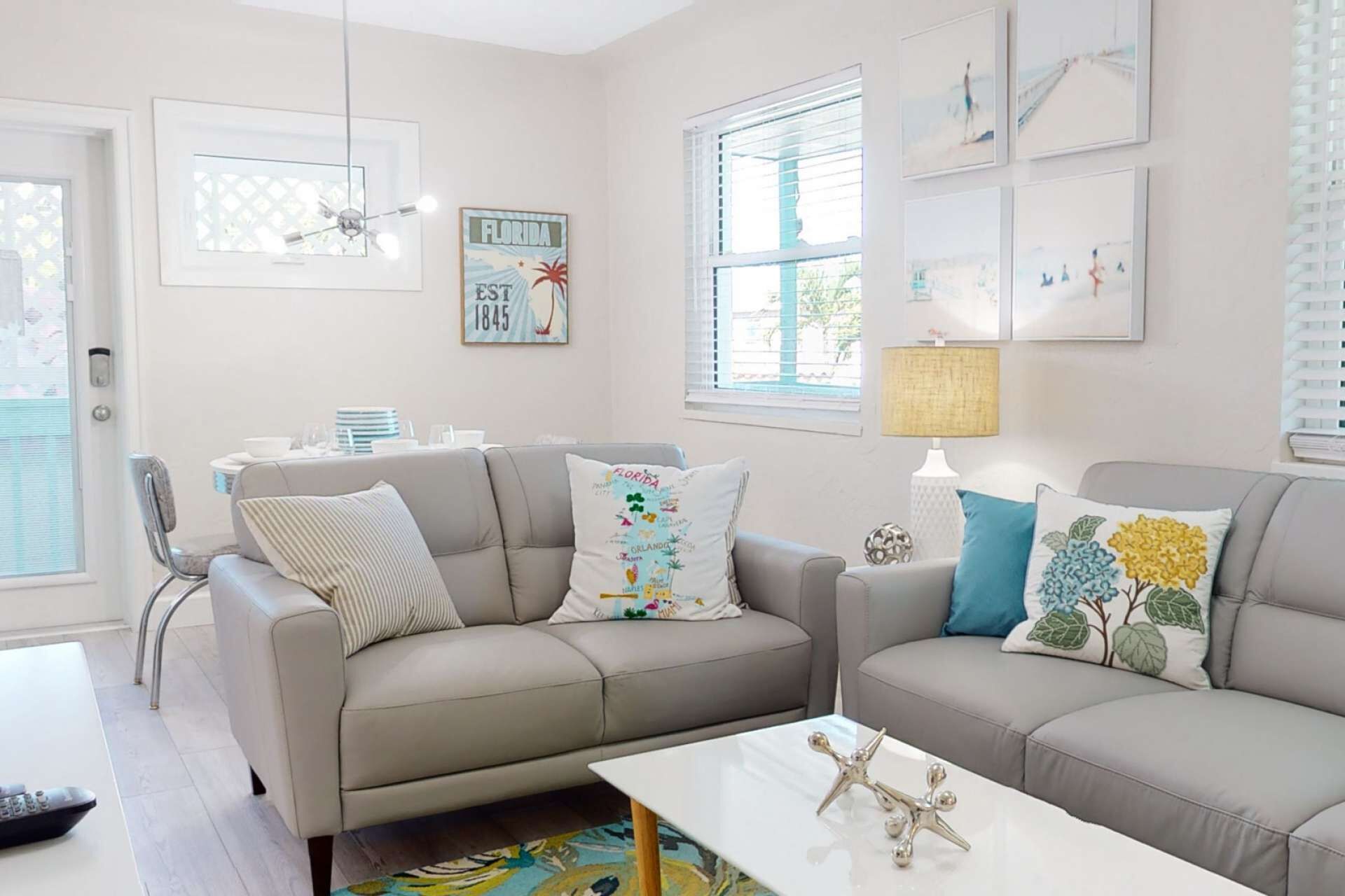 A living room with two grey sofas, a white coffee table, and a colorful rug. There are framed pictures on the walls and a lamp on the coffee table.