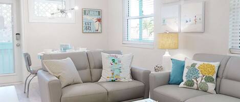 A living room with two grey sofas, a white coffee table, and a colorful rug. There are framed pictures on the walls and a lamp on the coffee table.