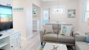 A living room with a TV, sofa, and coffee table. There is a door leading to another room, and a window with blinds.