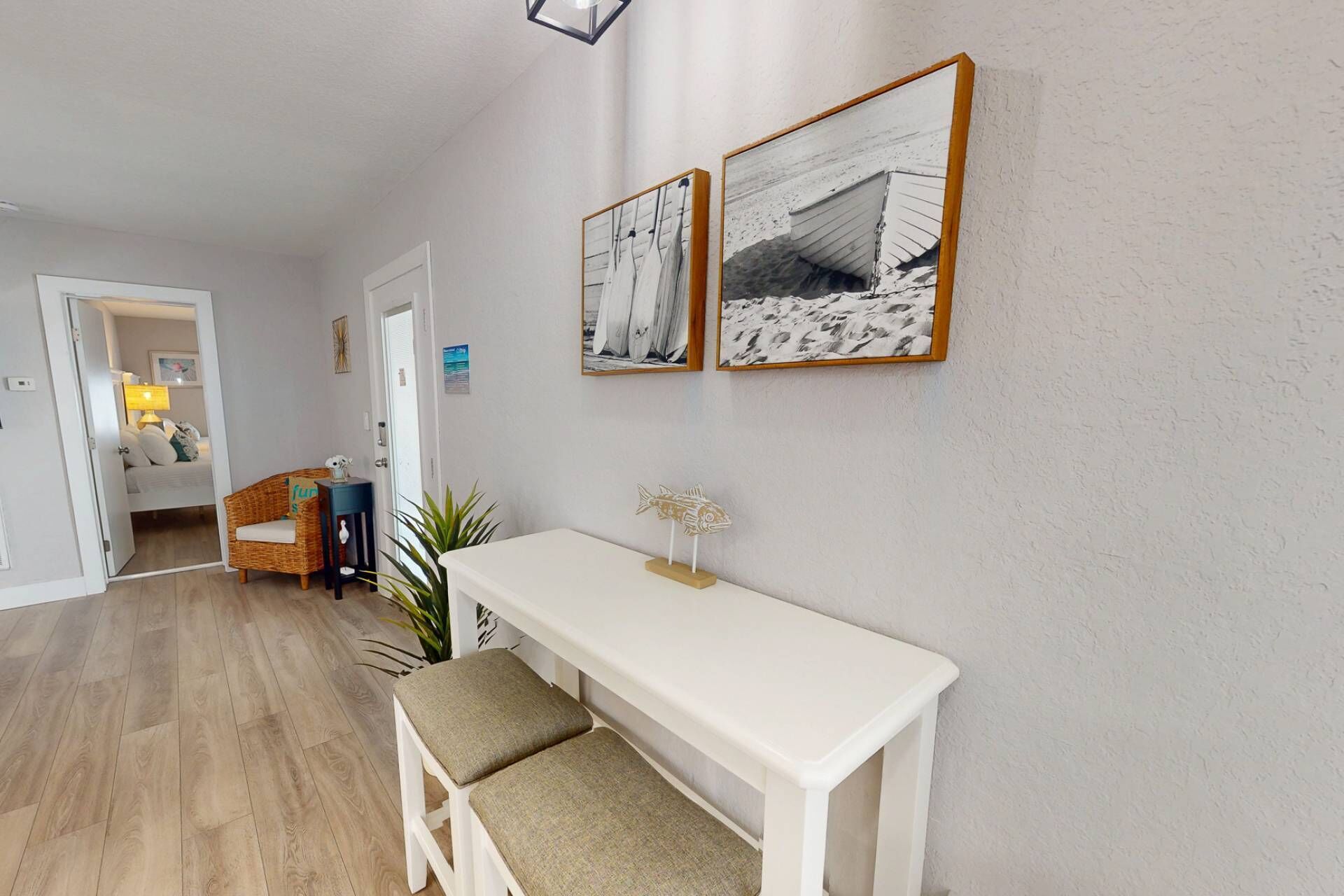 En hall med en vit bänk, en flätad stol och ett litet bord med en lampa. På väggen hänger två inramade tavlor.