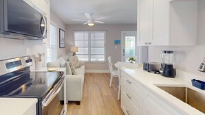 A compact kitchen with a stove, microwave, and a blender on the counter. There is a white sofa and a dining table with chairs. - Walk to Beach & Johns Pass Village ~ Heated Community Pool ~ Bikes ~ Paddle Boards/Kayaks (Madeira Beach)