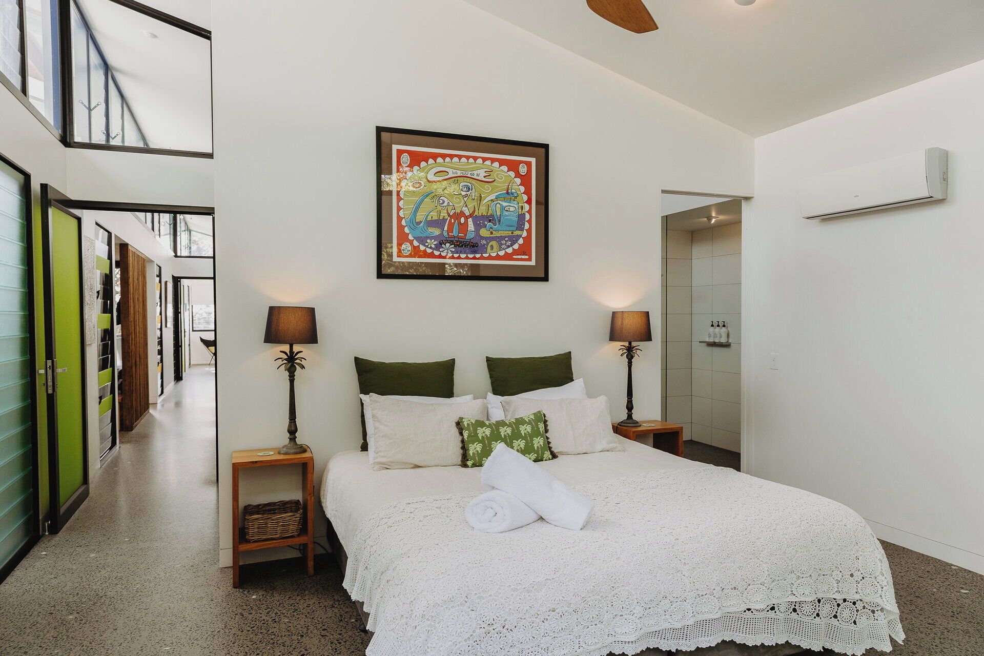 A bedroom with a bed, two bedside tables, a wall-mounted air conditioner, a framed artwork, and a hallway with a green door.