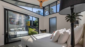A bedroom with a large bed, a nightstand, a lamp, and a view of a balcony with a chair and plants.