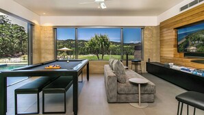 Un salon moderne doté d’une table de billard, de sièges confortables, d’un grand téléviseur et offrant une vue sur un jardin aménagé.
