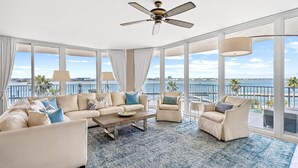 A spacious living room with a large sofa, a coffee table, and two armchairs. The room has large windows offering a view of the sea and palm trees. - Luxury Resort Spectacular Views of Cascading Pools and Bay; New Lazy River! Boat Slip! (Orange Beach)