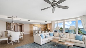 A spacious living area with a large sofa, dining table, and chairs, a kitchen in the background, and a balcony with a view. - Luxury Resort Spectacular Views of Cascading Pools and Bay; New Lazy River! Boat Slip! (Orange Beach)