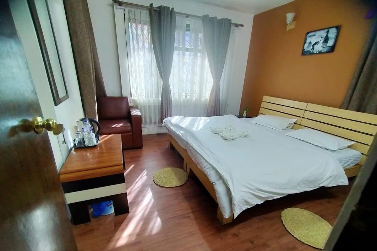 A hotel room with a wooden bed, a small desk with toiletries, a chair, and a view of the outside through a window with curtains.