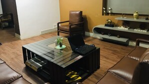 A living room with a glass coffee table, a brown leather chair, a sofa, a bookshelf with decorative items, and a framed picture on the wall.