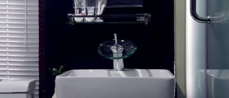 A bathroom with a white sink, a glass soap dish, and a shelf above the sink holding a toothbrush, toothpaste, and a glass.