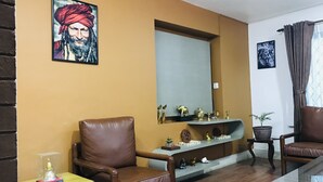 A living room with a brown leather sofa, a glass coffee table, and a wooden side table. There is a shelf with decorative items and a framed picture on the wall.