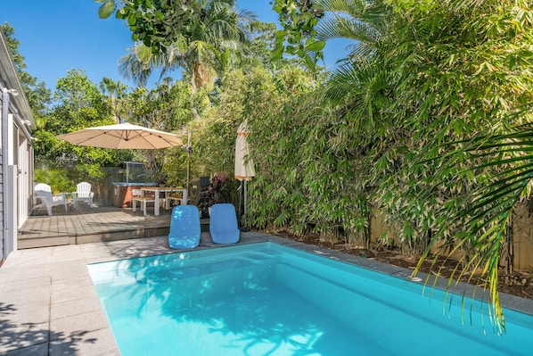 A swimming pool with two lounge chairs, a patio area with a table and chairs, and a lush green hedge surrounding the space.
