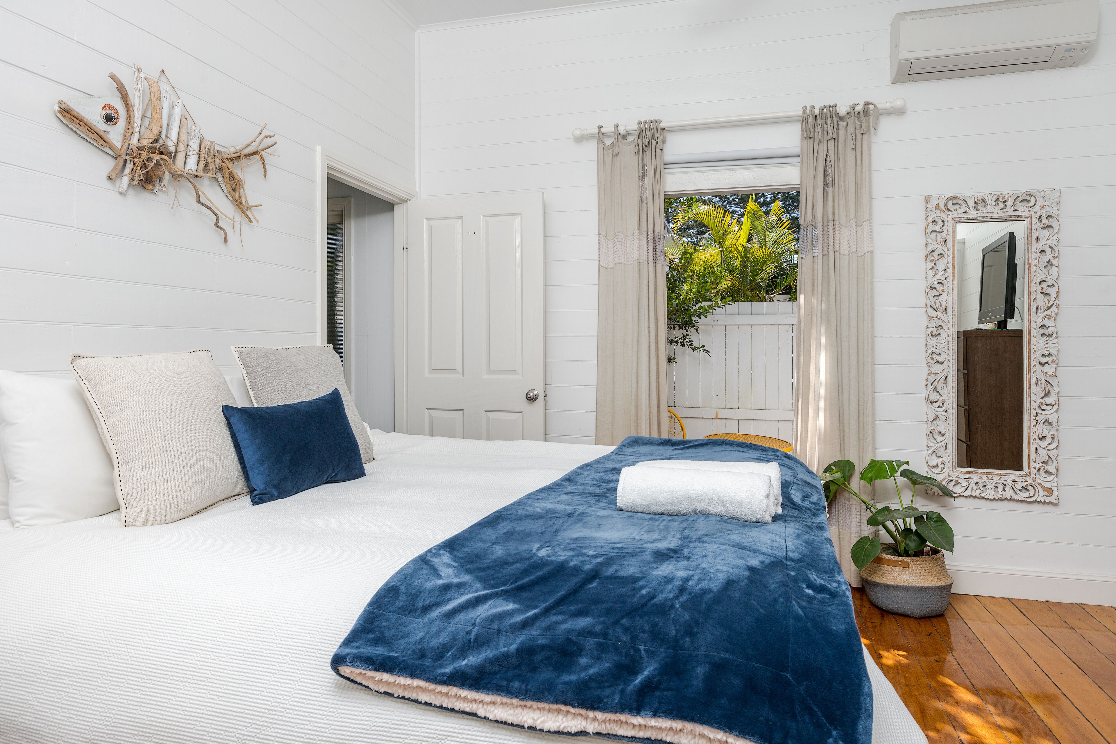 Quarto com cama grande, paredes brancas, arte de parede decorativa com peixes, espelho e planta em vaso.