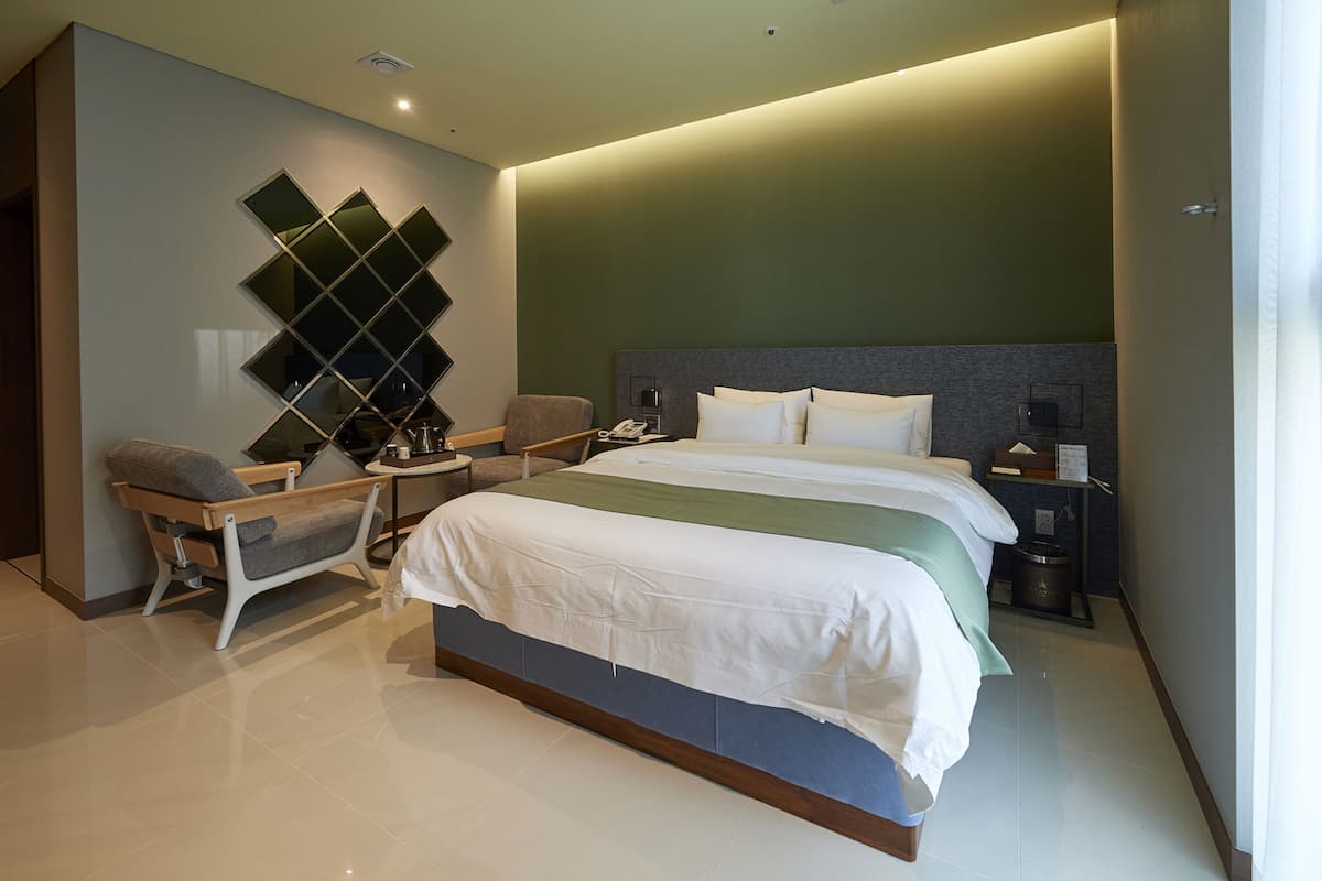 A modern hotel room with a large bed, a seating area with a chair, and a wall-mounted mirror.