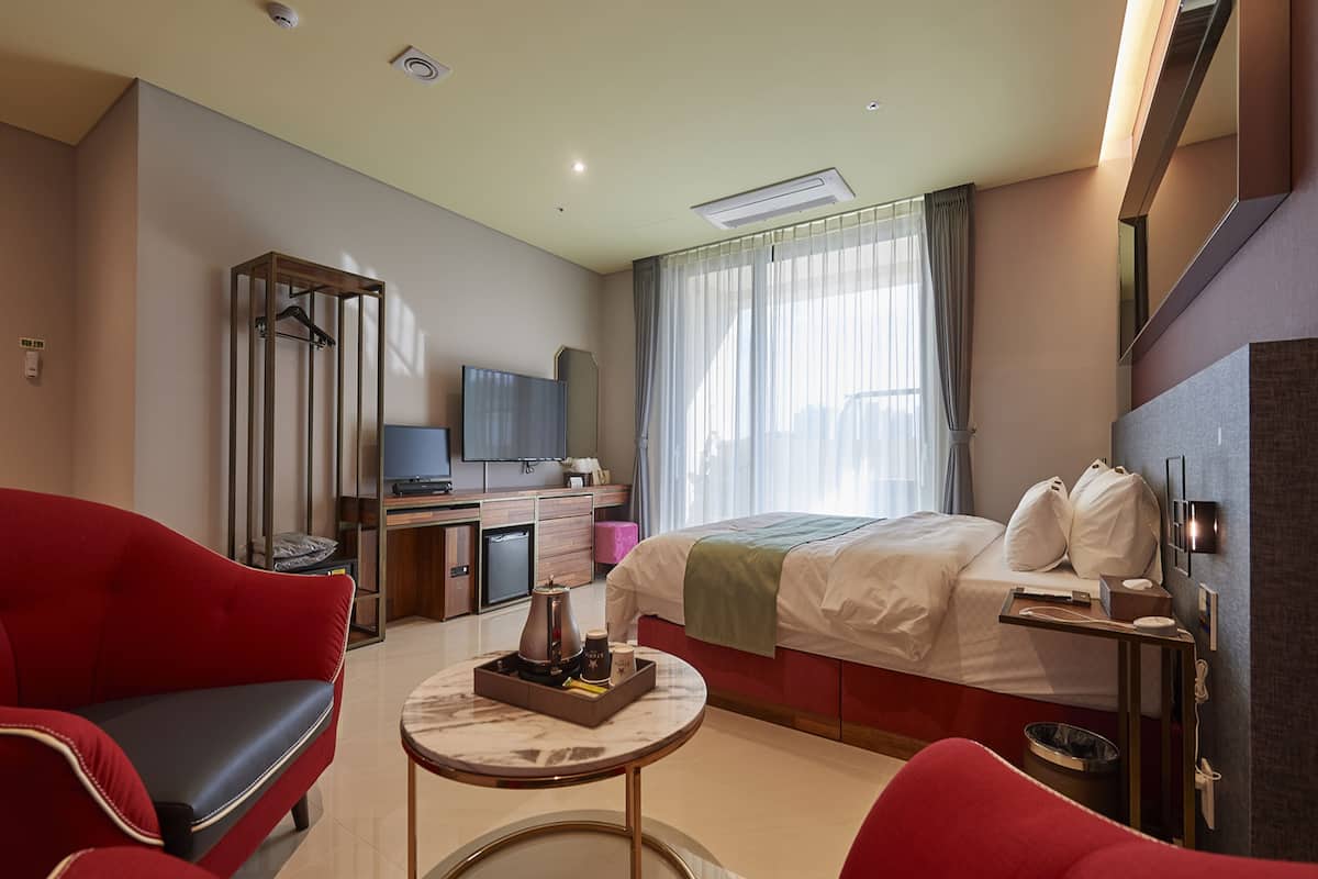 A hotel room with a bed, a desk with a TV, a red armchair, a coffee table, and a minibar.