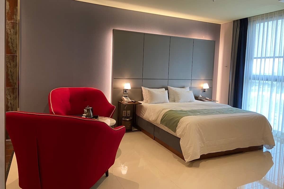 A modern hotel room with a large bed, a red armchair, a bedside table with a lamp, and a view of the outdoors through large windows.