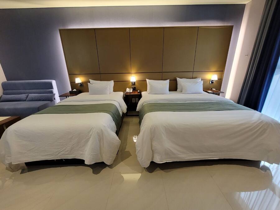 Una camera d'albergo con due letti, una poltrona blu, comodini con lampade e una testiera grande.
