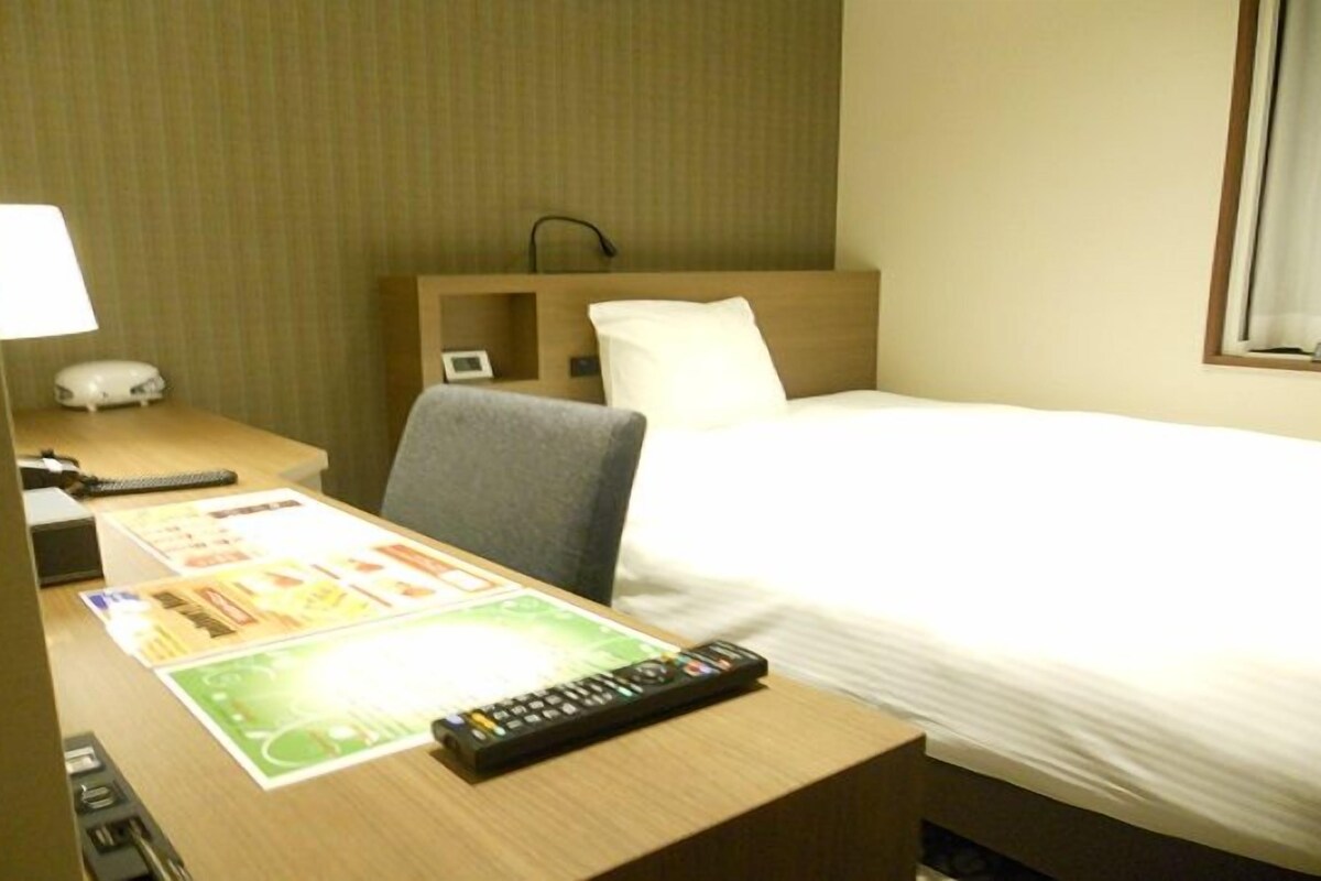 A hotel room with a bed, a desk with a remote control, a lamp, and a wall-mounted phone.