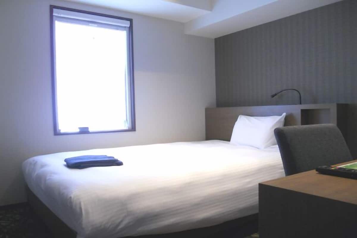 A hotel room with a bed, a desk, a chair, a window, and a wall-mounted lamp.