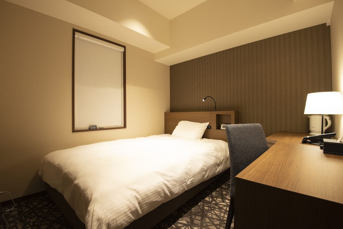 A hotel room with a bed, a desk, a chair, a lamp, and a wall-mounted TV.