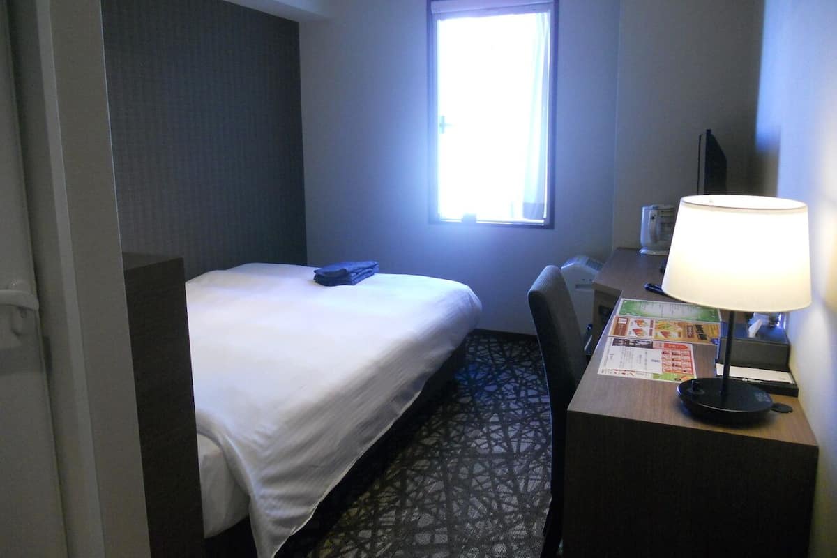 A hotel room with a bed, a desk with a lamp, a window, and a wall-mounted TV.