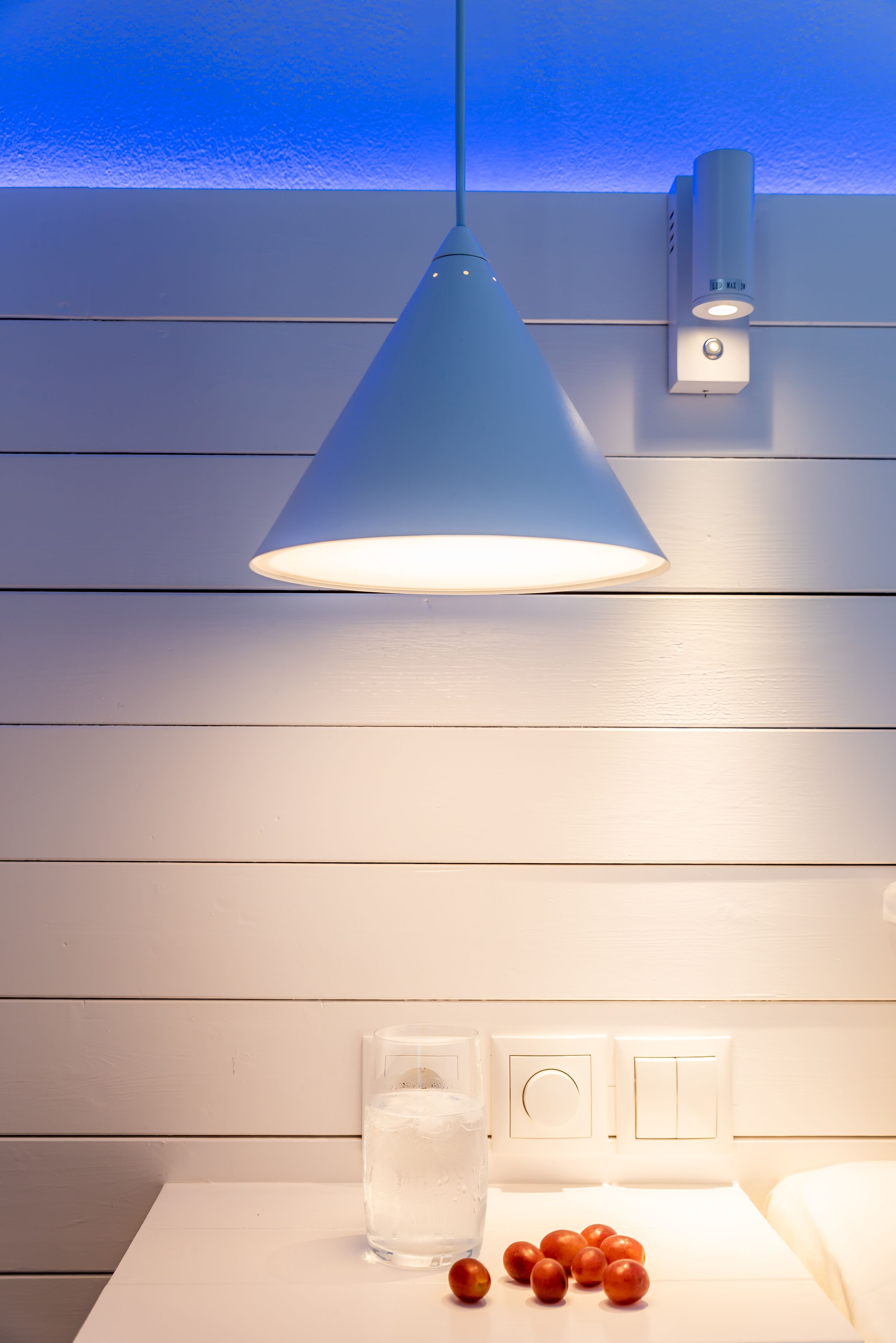 Ett rum med en vit vägg, en blå hängande lampa, ett glas vatten och några äpplen på ett bord.